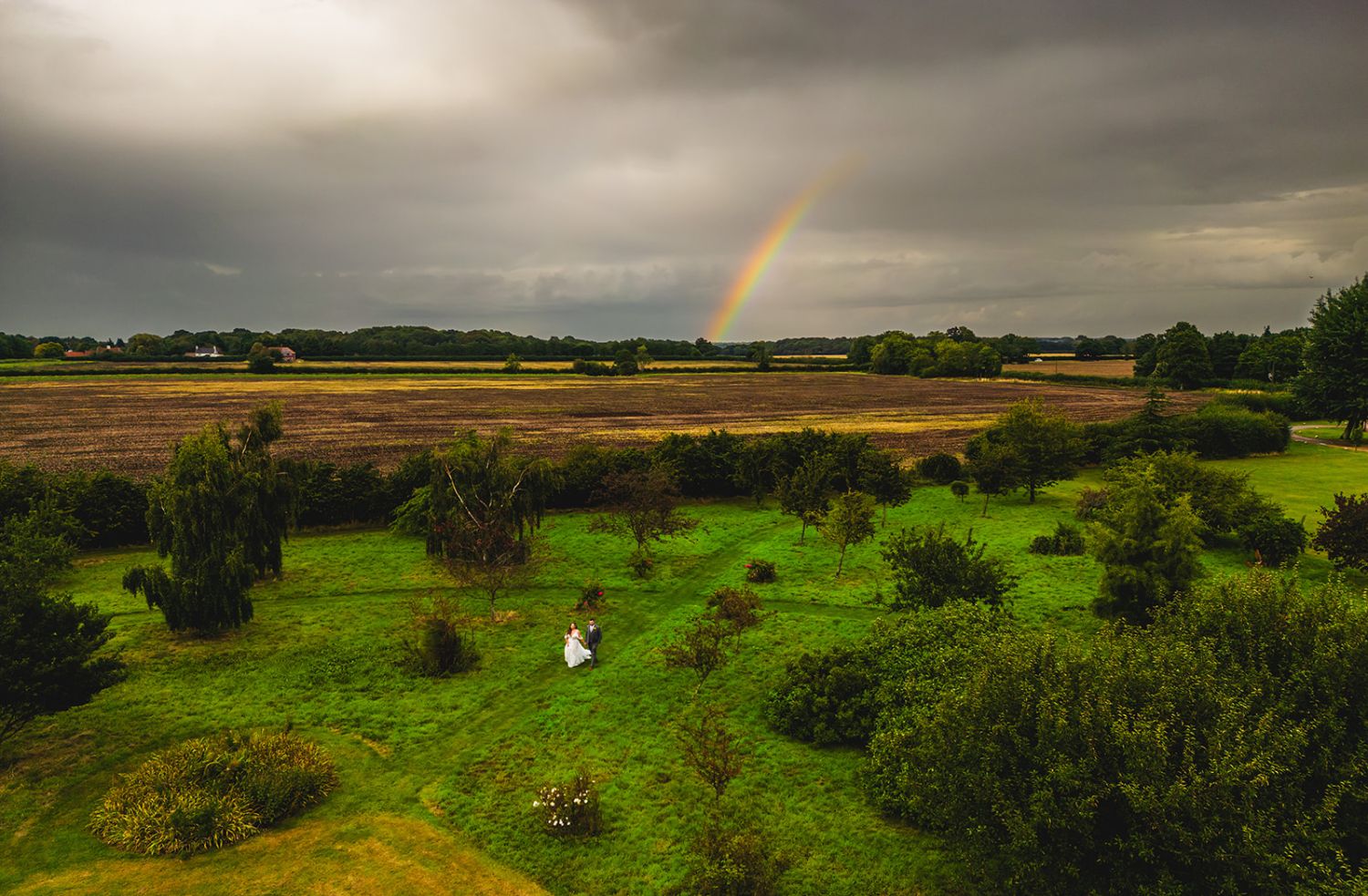 bride and groom walking through a field with a rainbow overhead